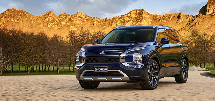 Blue Mitsubishi Outlander on paved road with mountains in the background